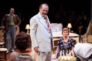 Several people gather around a birthday cake with lit candles. The scene suggests a family celebration, with attention focused on the cake and the person being celebrated.