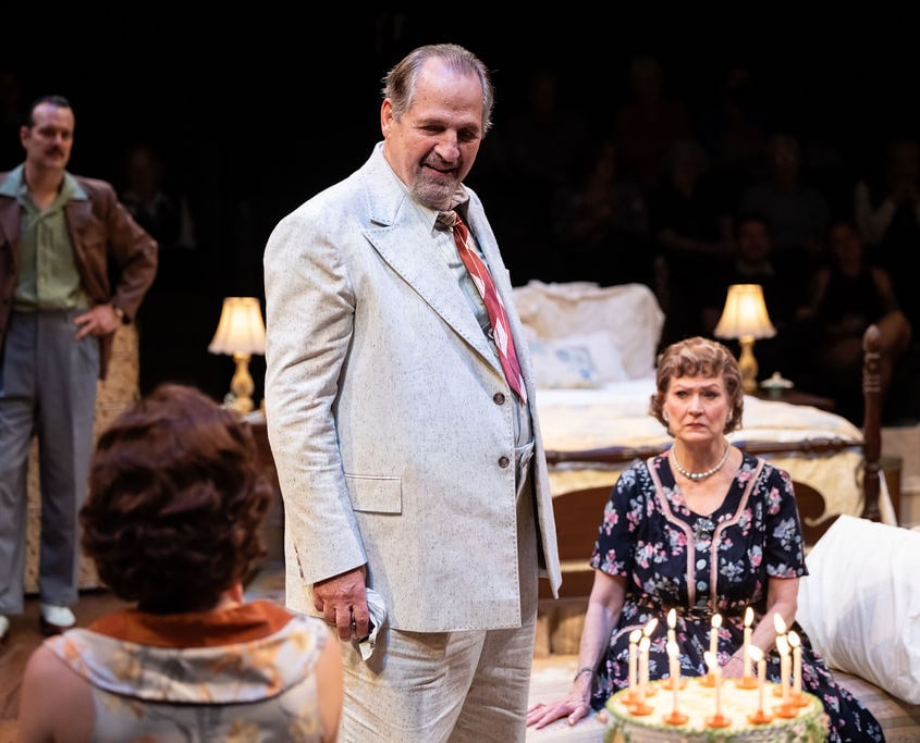 Several people gather around a birthday cake with lit candles. The scene suggests a family celebration, with attention focused on the cake and the person being celebrated.