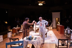 Two men raise glasses in a toast, sharing a moment of camaraderie. The room’s vintage decor and warm lighting add to the celebratory atmosphere.