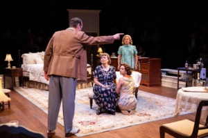 A dramatic scene unfolds as one person points accusingly at another kneeling beside an older seated individual. The setting is a richly furnished bedroom, and the interaction appears emotionally charged.