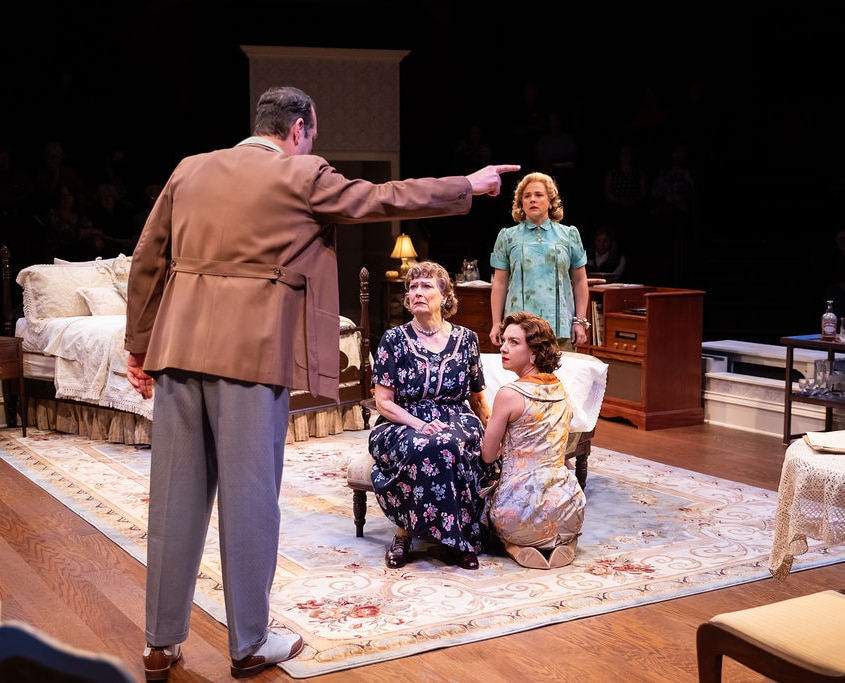 A dramatic scene unfolds as one person points accusingly at another kneeling beside an older seated individual. The setting is a richly furnished bedroom, and the interaction appears emotionally charged.