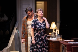A distressed individual is comforted by another in a close, emotional exchange. The vintage bedroom setting enhances the intimacy and gravity of the moment.