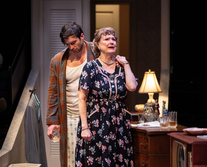 A distressed individual is comforted by another in a close, emotional exchange. The vintage bedroom setting enhances the intimacy and gravity of the moment.