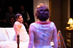 A person sits upright in bed, gazing toward another individual standing nearby. The vintage bedroom setting and their expressions suggest a moment of confrontation or revelation.