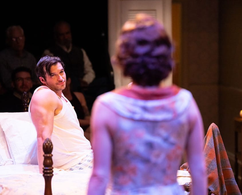 A person sits upright in bed, gazing toward another individual standing nearby. The vintage bedroom setting and their expressions suggest a moment of confrontation or revelation.