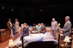 A group of people gather in a bedroom decorated with vintage furniture and warm lighting. The arrangement and expressions suggest a serious or reflective moment among family members.