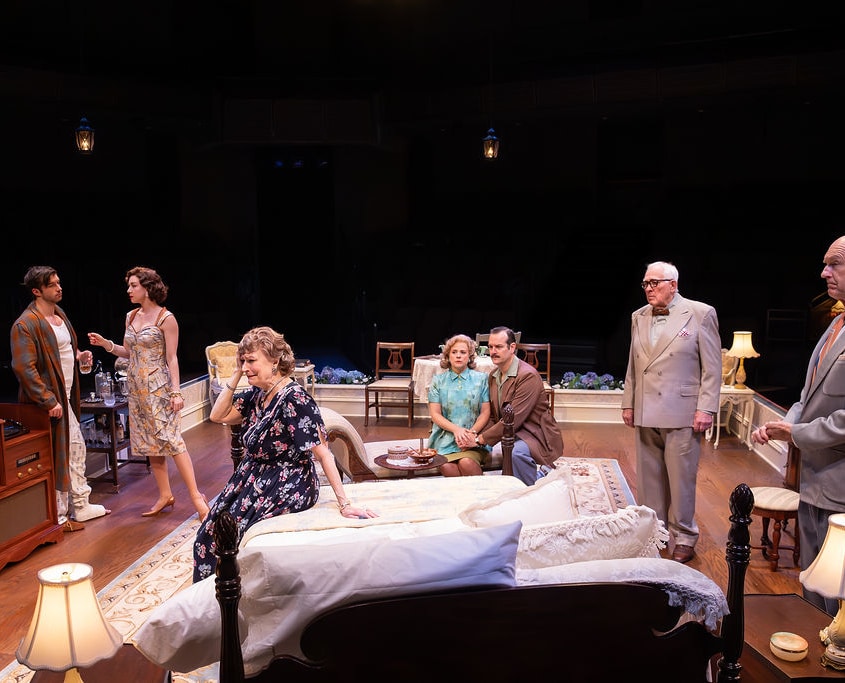 A group of people gather in a bedroom decorated with vintage furniture and warm lighting. The arrangement and expressions suggest a serious or reflective moment among family members.