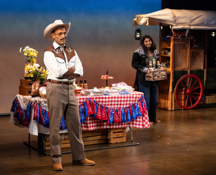 Rajah Sen stands confidently in Western attire with arms crossed beside a festive table adorned with red checkered cloth and decorations. His daughter, Lakshmi Sen, smiles in the background, holding a crate near a vintage wagon.