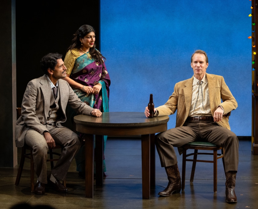 Richard Larson sits at a table dressed in a tan suit and a bolo tie, beer bottle in hand. Rajah sits listening at the table dressed in a grey suit while Chitra Sen stands behind him dressed in a traditional Indian sari in burgundy and teal with gold details.