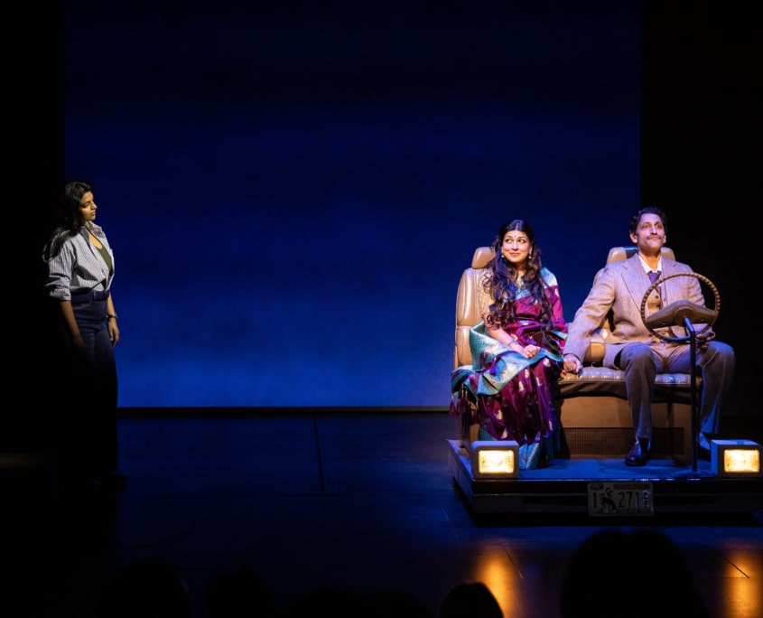 Lakshmi Sen looks on at her parents from a distance while they sit in a car holding hands with soft smiles on their face. Chitra Sen wears a traditional Indian sari in burgundy and teal with gold details while Rajah Sen wears a grey suit.