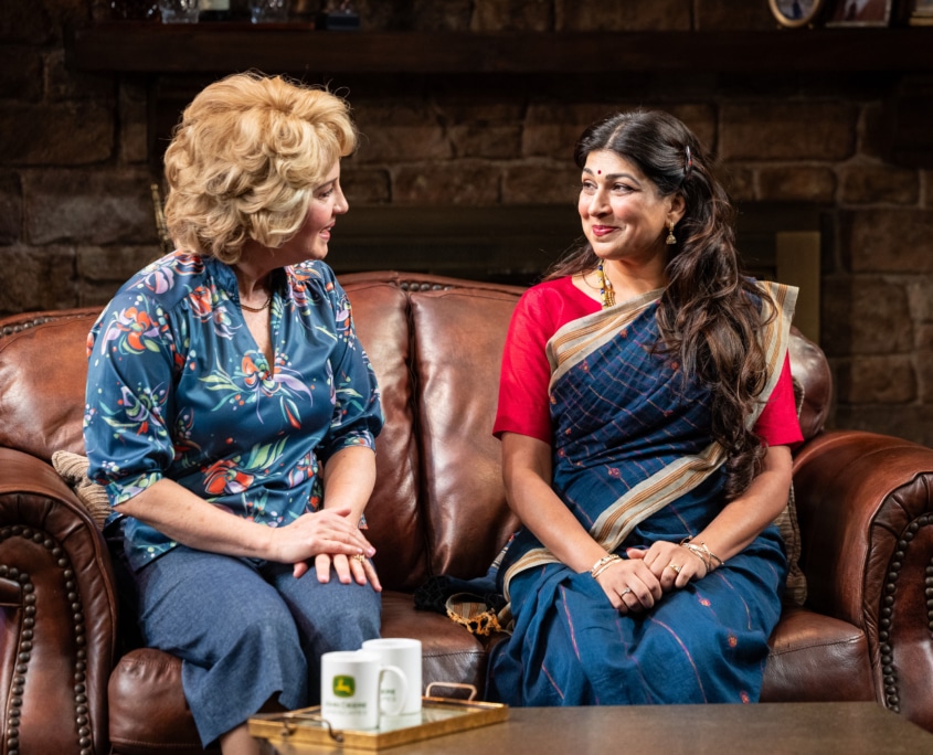 Nancy Larson and Chitra Sen sit on a brown leather couch in a cozy room. Nancy wears a floral blouse and jeans while Chitra wears a traditional Indian sari in red and blue with tan details. They share a warm, friendly conversation.