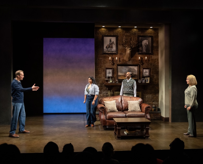 Richard Larson gestures toward his Western style living room with photos of horses and a taxidermized deer on the wall. Rajah Sen and his daughter, Laksmi, listen along with Nancy Larson.