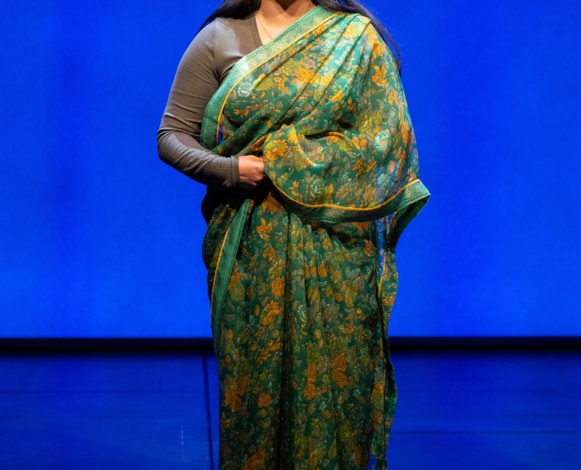 Laksmi Sen stands smiling in a traditional Indian sari in green and yellow with Western-style boots on.