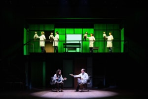On a two-level stage, several people in white lab coats move across the upper platform holding small dolls, while a woman and a man in a doctor’s coat sit facing each other in roll on the lower level.