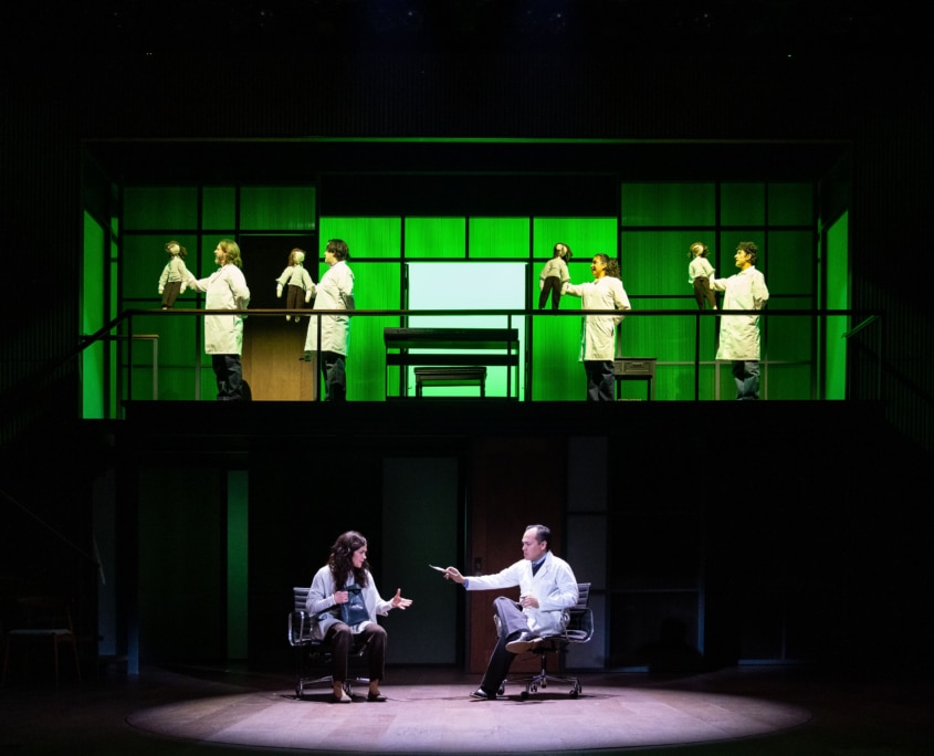 On a two-level stage, several people in white lab coats move across the upper platform holding small dolls, while a woman and a man in a doctor’s coat sit facing each other in roll on the lower level.