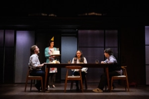 Three people sit around a wooden table while a woman wearing a party hat stands next to the table holding a cake with lit candles.