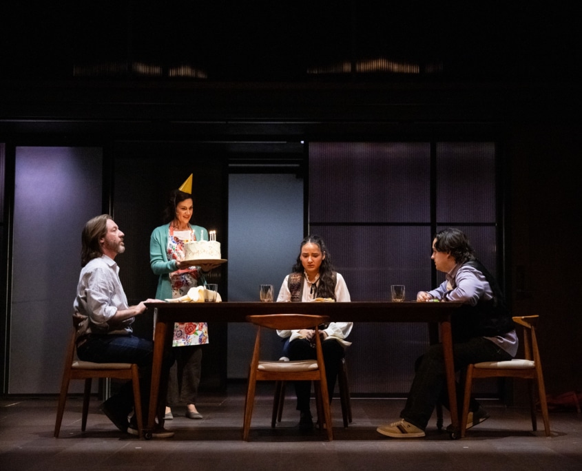 Three people sit around a wooden table while a woman wearing a party hat stands next to the table holding a cake with lit candles.