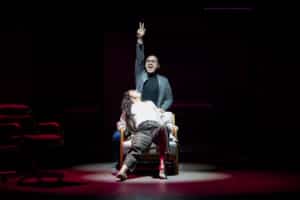 A woman reclines in a chair at center stage while a man wearing glasses stands behind the chair, pointing upward with one hand. A pool of red-tinted light surrounds the chair, with darkness filling the rest of the stage.