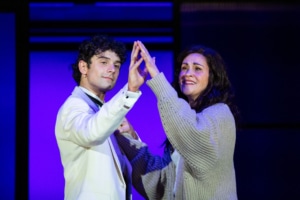 A young man and his mother stand close together on stage, raising their hands to touch palm to palm. The background features purple lighting with rectangular set panels.