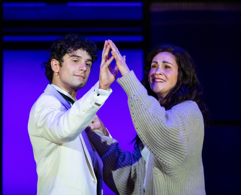 A young man and his mother stand close together on stage, raising their hands to touch palm to palm. The background features purple lighting with rectangular set panels.