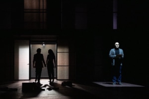 A dark stage with a doorway at left where two silhouetted figures walk out hand in hand, while another person stands alone at right on a lit platform holding papers.