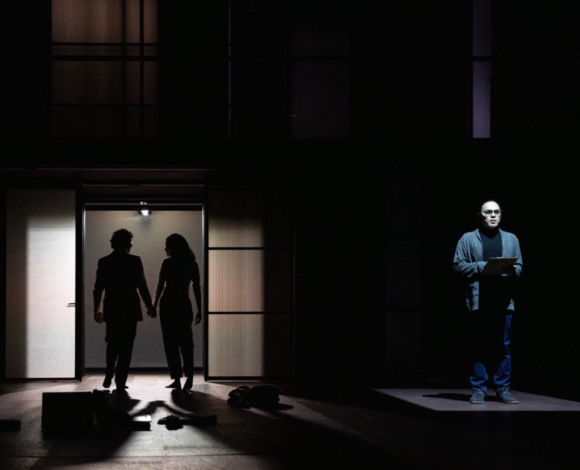 A dark stage with a doorway at left where two silhouetted figures walk out hand in hand, while another person stands alone at right on a lit platform holding papers.