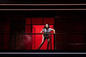 A girl stands on an elevated walkway, leaning forward on a railing. A bright red doorway and red wall panels fill the background.