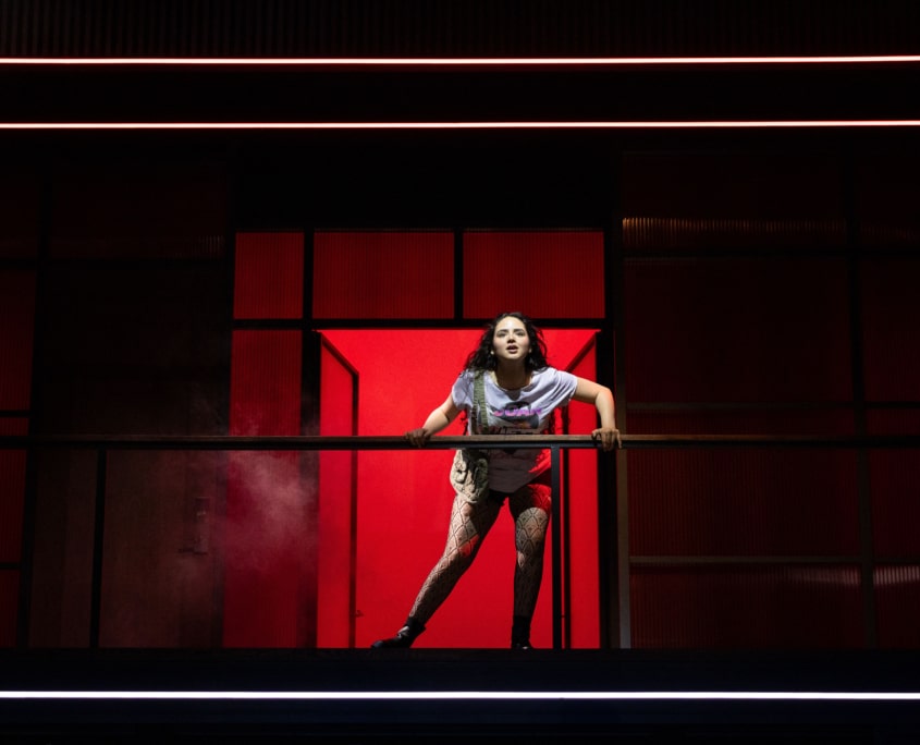 A girl stands on an elevated walkway, leaning forward on a railing. A bright red doorway and red wall panels fill the background.