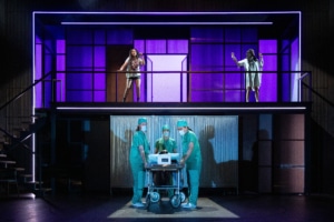 Two women stand on an upper balcony with purple lit panels behind them, while three people in medical scrubs gather around a wheeled hospital gurney on the lower level.
