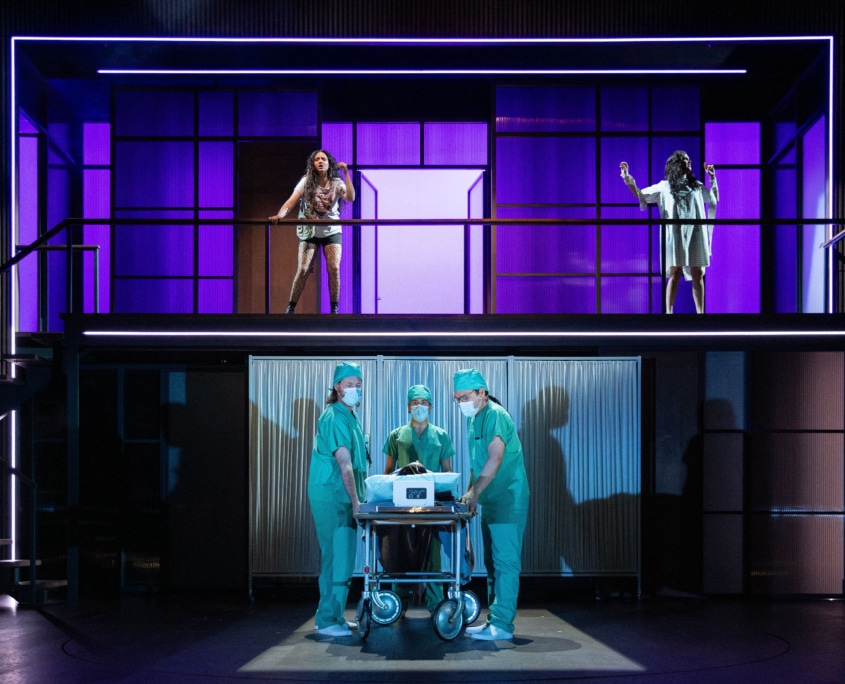 Two women stand on an upper balcony with purple lit panels behind them, while three people in medical scrubs gather around a wheeled hospital gurney on the lower level.