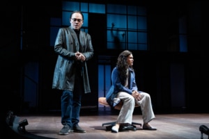 A man in glasses and a long dark coat stands to the left, while a woman in jeans and an blazer sits on a chair at right. A large window-like grid fills the background, with a shadowed hand pressed against the glass.