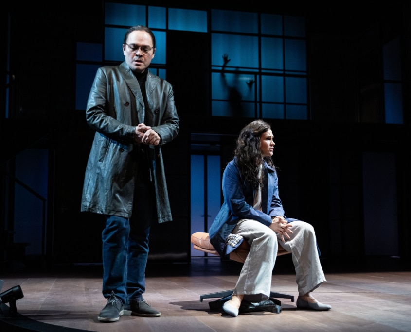 A man in glasses and a long dark coat stands to the left, while a woman in jeans and an blazer sits on a chair at right. A large window-like grid fills the background, with a shadowed hand pressed against the glass.