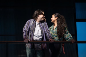 A young man and woman lean toward each other across a railing on an elevated platform, gazing affectionately at one another. The background is dark with vertical blue-lit panels.