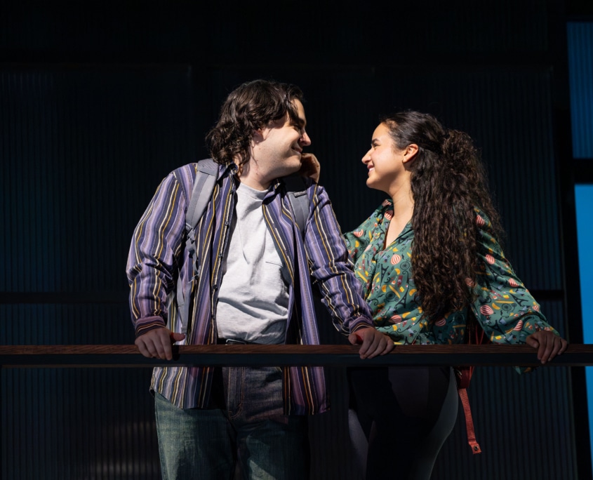 A young man and woman lean toward each other across a railing on an elevated platform, gazing affectionately at one another. The background is dark with vertical blue-lit panels.