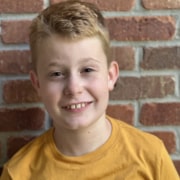 Headshot of young actor with cropped redish hair, full grin, and yellow shirt in front of brick wall.