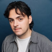 Headshot of an actor on a dark blue background wearing a white t-shirt with a gray shirt and dark hair.