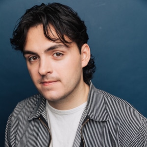 Headshot of an actor on a dark blue background wearing a white t-shirt with a gray shirt and dark hair.