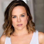 Headshot of actress with wavy brown hair, blue eyes, and white sleeveless top.