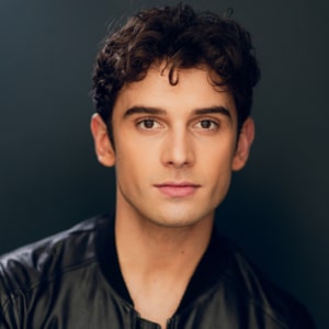 Headshot of an actor on a dark gray background wearing a black shirt with short dark hair.