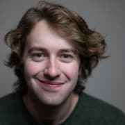 Headshot of an actor with gray background, black t-shirt, wavy short brown hair, and a grin.