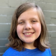 Headshot of young actor with shoulder-length brown hair swept to one side, large grin, and bright blue crew-neck t-shirt in front of gray brick wall.