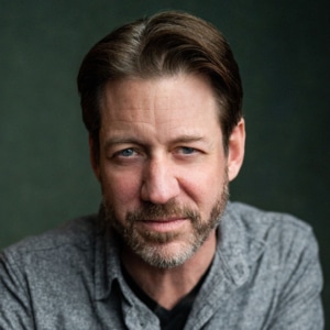 Headshot of an actor on a dark backgoun wearing a gray shirt with brown hair and a beard.