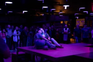 A dimly lit room with purple lighting, featuring a group of people standing and observing two women sitting on a raised platform in the center. The women on the platform are seated close together, with one leaning against the other. The background shows more people standing and watching, as well as some shelving and light fixtures.
