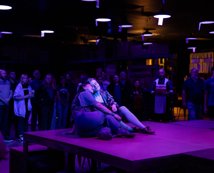 A dimly lit room with purple lighting, featuring a group of people standing and observing two women sitting on a raised platform in the center. The women on the platform are seated close together, with one leaning against the other. The background shows more people standing and watching, as well as some shelving and light fixtures.