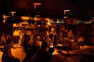 A dimly lit room resembling a warehouse, with shelves filled with cardboard boxes. There are people seated around tables. Hanging lights illuminate the space. A person stands near the center places a box on a shelf in the center of the room.