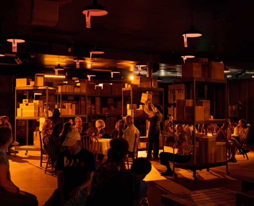 A dimly lit room resembling a warehouse, with shelves filled with cardboard boxes. There are people seated around tables. Hanging lights illuminate the space. A person stands near the center places a box on a shelf in the center of the room.