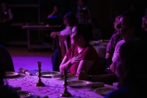 A dimly lit room with a purple hue, where several people are seated at a table set for a meal. The table is covered with a white cloth and features plates, napkins, and two candlesticks.