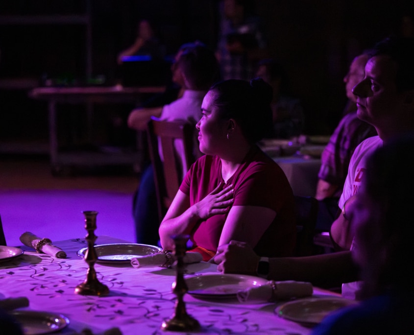 A dimly lit room with a purple hue, where several people are seated at a table set for a meal. The table is covered with a white cloth and features plates, napkins, and two candlesticks.