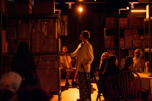 A dimly lit room with shelves full of cardboard boxes. One person stands near the shelves, observing the items on the shelf. The room is bathed in a warm, orange glow, creating a cozy atmosphere.