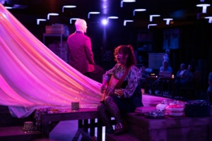 A dimly lit scene with purple and pink lighting, featuring a woman seated on a wooden platform playing an acoustic guitar. Another individual in a suit sits nearby, facing away. Decorative items and casserole dishes are arranged around the platform, while the audience observes the performance.